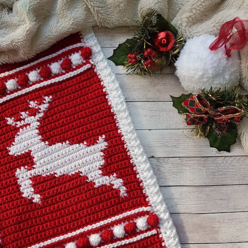 May include: A red and white crocheted Christmas stocking with a white reindeer design and white pom-poms around the edges.