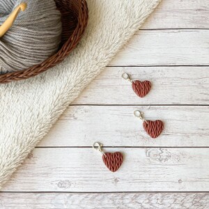 May include: Three heart-shaped stitch markers in a reddish-brown color with silver clasps. A wooden crochet hook and a ball of gray yarn are in a brown woven basket. The background is a white, fluffy fabric and a light wood surface.