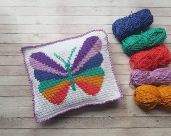 Op de afbeelding: Een gehaakt kussen met een kleurrijk vlinderontwerp. Het kussen is wit met een paarse, groene, oranje en roze vlinder in het midden. Er liggen zes bollen garen in verschillende kleuren naast het kussen.