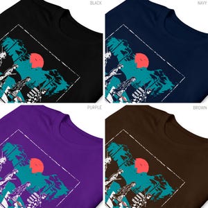 May include: Four t-shirts in black, navy, purple, and brown, each featuring a graphic design. The design includes a red sun, teal trees, and figures in a white outlined square. The shirts are made of a soft material.