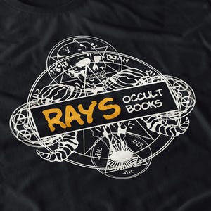 May include: Black t-shirt featuring a white occult design with a skull, geometric shapes, and the words "RAYS OCCULT BOOKS" in orange and white. The design is centered on the shirt.