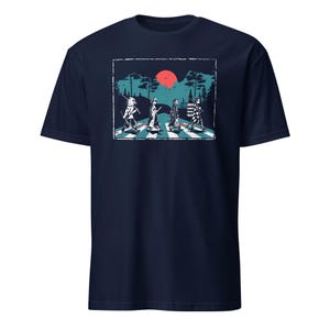 May include: Navy blue t-shirt featuring a graphic of four figures crossing a crosswalk, with a red sun and trees in the background. The figures are in a line, with a white and black crosswalk. The text on the shirt is in white.