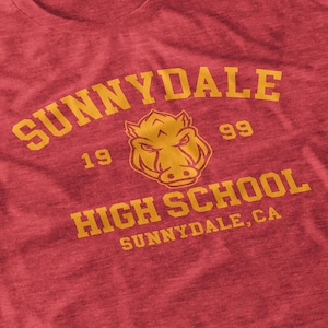 May include: Red t-shirt with gold text reading "SUNNYDALE HIGH SCHOOL". The shirt features a gold illustration of a wild boar's head. The year "1999" is printed above the boar's head. The text "SUNNYDALE, CA" is also visible.