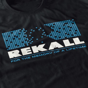 May include: Black t-shirt with the word "REKALL" in large white letters. Above the word is a blue pixelated design. Below the word is the text "FOR THE MEMORY OF A LIFETIME" in blue.