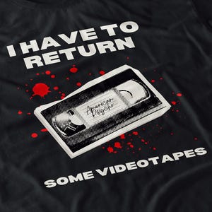 May include: Black t-shirt with the text "I HAVE TO RETURN SOME VIDEOTAPES." A graphic of a VHS tape with "American Psycho" written on it is in the center, with red splatter details.