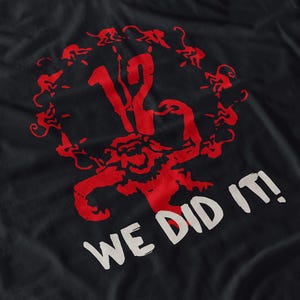 May include: Black t-shirt with a red graphic. The graphic features the number 12, surrounded by a circle of monkey-like figures, and a central figure. The words "WE DID IT!" are printed in white.