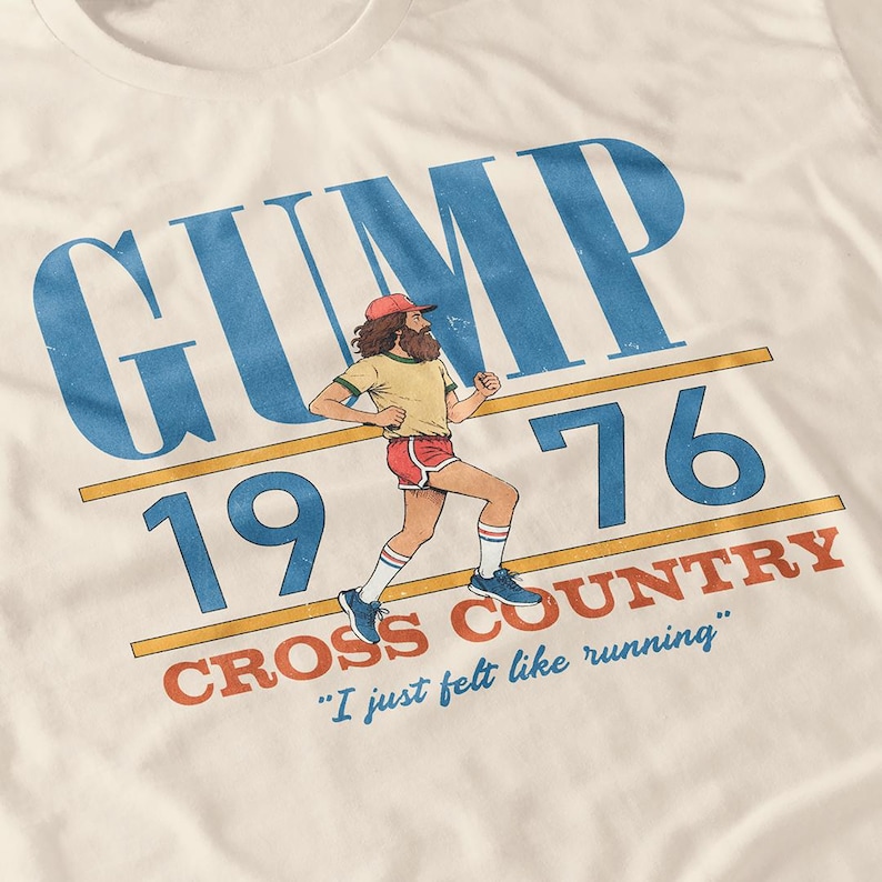 Puede incluir: Una camiseta de color crema con un gr&aacute;fico de un corredor barbudo con gorra roja, camiseta amarilla y pantalones cortos rojos. El texto dice GUMP 19 76 CROSS COUNTRY e I just felt like running en letras azules y rojas con estilo vintage.