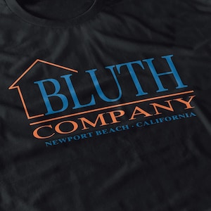 Puede incluir: Camiseta negra con el texto "BLUTH COMPANY" en azul y naranja, con un contorno de casa encima de la palabra "BLUTH". El texto "NEWPORT BEACH - CALIFORNIA" está debajo.