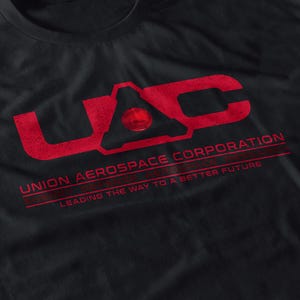 May include: Black t-shirt featuring a distressed red graphic of the letters "UAC" with the words "UNION AEROSPACE CORPORATION" and "LEADING THE WAY TO A BETTER FUTURE" printed below. The shirt is made of a soft material.