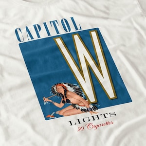 May include: White t-shirt featuring a vintage advertisement design. The design includes the text "CAPITOL" in blue, a large white and gold "W" on a blue background, and an illustration of a woman in Native American attire. The text "LIGHTS 20 Cigarettes" is also present.