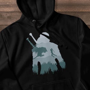 May include: Black hooded sweatshirt featuring a silhouette design of a warrior, a sword, and a mythical creature against a backdrop of mountains and a forest. The graphic is in shades of blue and gray.