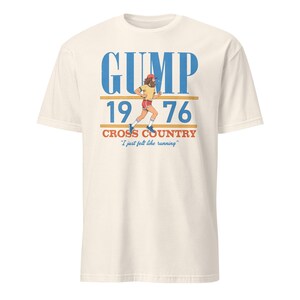 Puede incluir: Una camiseta color hueso con un gr&aacute;fico de un corredor con barba y gorra roja. El texto dice GUMP 1976 CROSS COUNTRY y I just felt like running en tinta azul, naranja y roja. El dise&ntilde;o incluye l&iacute;neas horizontales por encima y por debajo del a&ntilde;o.