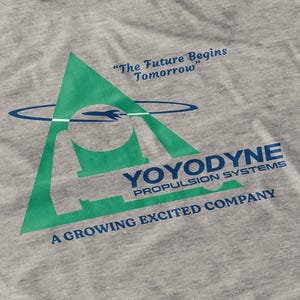 Puede incluir: Camiseta gris con un diseño de triángulo verde, un cohete y el texto "YOYODYNE Propulsion Systems". La camiseta también incluye las frases "The Future Begins Tomorrow" y "A Growing Excited Company".
