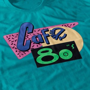 May include: Teal t-shirt with a retro graphic design. The design features the word "Cafe" in blue, a yellow circle, a pink diamond shape with black speckles, and the number "80" in green on a black rectangle.