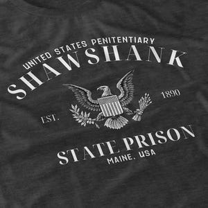 May include: Dark gray t-shirt featuring a graphic design with the text "SHAWSHANK" and "UNITED STATES PENITENTIARY" in a curved font. An eagle with outstretched wings and a shield is in the center, with "STATE PRISON MAINE, USA" below.