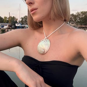 Può includere: Collana con pendente a forma di conchiglia color argento e cordino bianco. Il pendente ovale presenta dettagli intagliati. La persona nell'immagine indossa un top nero senza spalline e pantaloni neri. Lo sfondo mostra acqua e barche.
