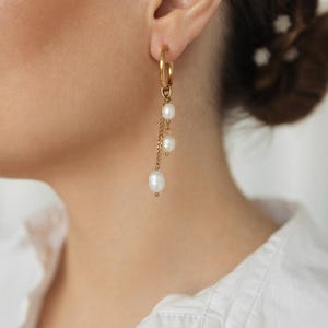 Puede incluir: Pendientes de aro dorados con tres perlas blancas colgantes. Los pendientes están sujetos a una delicada cadena dorada. Una persona lleva una camisa blanca.