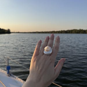 May include: A ring featuring a large, smooth, white seashell centrepiece with a spiralled, pearlescent top. The shell is mounted on a simple, polished silver-toned band that sits prominently on the finger, showcasing a natural, organic aesthetic.