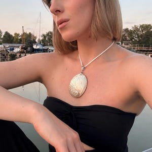 Peut inclure: Un collier avec un pendentif argenté de forme ovale et texturé. Le pendentif est suspendu à un cordon blanc. La personne porte un haut noir sans bretelles et un pantalon noir. L'arrière-plan montre un plan d'eau et des bateaux.