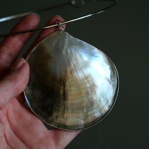 May include: A necklace featuring a large, iridescent mother-of-pearl shell pendant with a metallic rim. The shell displays natural swirling patterns in shades of cream, brown, and gray. It is suspended from a thin, circular silver-toned metal collar.