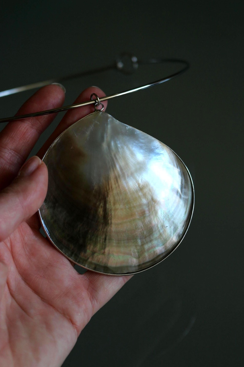 May include: A necklace featuring a large, iridescent mother-of-pearl shell pendant with a metallic rim. The shell displays natural swirling patterns in shades of cream, brown, and gray. It is suspended from a thin, circular silver-toned metal collar.