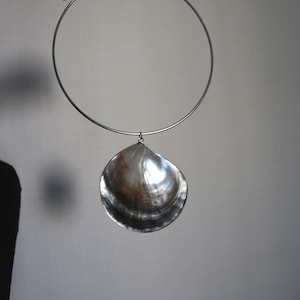 May include: A silver-toned metal choker necklace featuring a single, large, iridescent seashell pendant. The shell displays a dark, glossy, and pearlescent surface with natural brown and grey color variations, suspended from a thin, circular metal band.