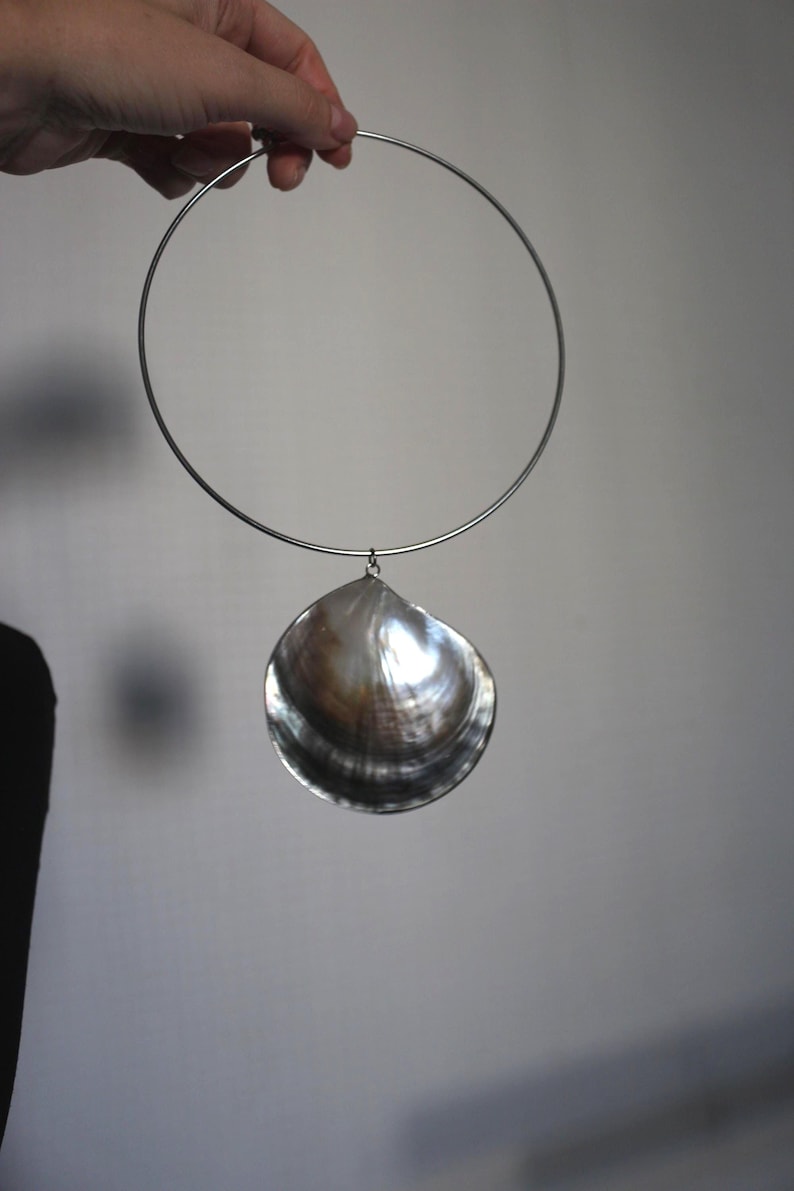 May include: A silver-toned metal choker necklace featuring a single, large, iridescent seashell pendant. The shell displays a dark, glossy, and pearlescent surface with natural brown and grey color variations, suspended from a thin, circular metal band.