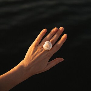May include: A ring featuring a polished, cream-colored spiral seashell as the central focal point. The shell has a smooth, rounded surface with a natural coiled structure, securely mounted on a band that rests on a finger, catching warm light.