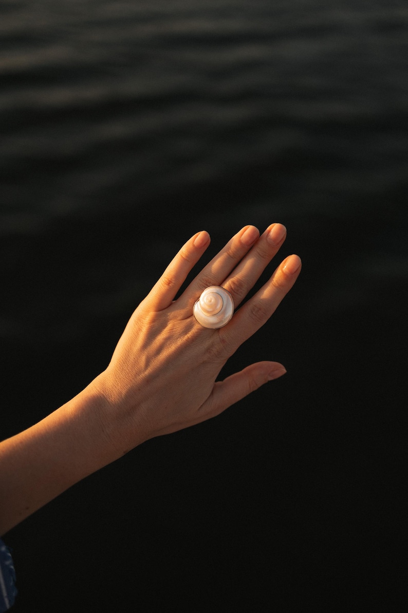 May include: A ring featuring a polished, cream-colored spiral seashell as the central focal point. The shell has a smooth, rounded surface with a natural coiled structure, securely mounted on a band that rests on a finger, catching warm light.