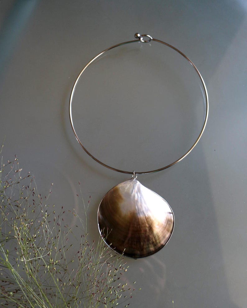 May include: A silver-toned metal choker necklace featuring a single, large, rounded seashell pendant. The shell displays a gradient of iridescent white and deep brown tones, suspended from a thin, circular wire band with a simple hook closure.