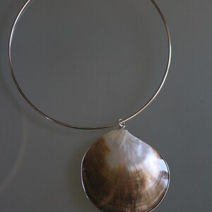 May include: A silver-toned metal choker necklace featuring a single, large, iridescent seashell pendant. The shell displays natural brown and white color gradients with a smooth, polished surface, suspended from a thin, circular wire collar.