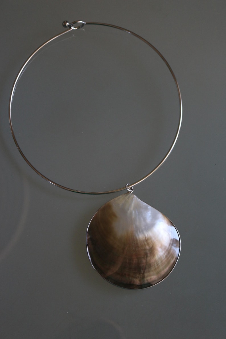 May include: A silver-toned metal choker necklace featuring a single, large, iridescent seashell pendant. The shell displays natural brown and white color gradients with a smooth, polished surface, suspended from a thin, circular wire collar.