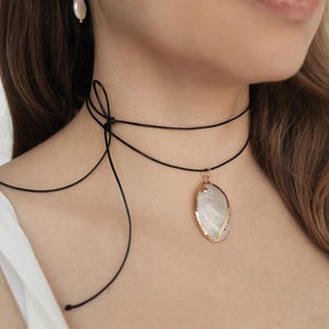 May include: A black choker necklace with a pear-shaped pendant featuring a white and gold design. A matching gold hoop earring with a white pearl hangs from the ear.