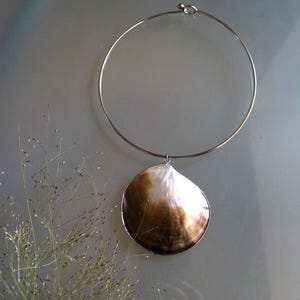 May include: A silver-toned metal choker necklace featuring a single, large, rounded seashell pendant. The shell displays a gradient of colors, transitioning from a deep, dark brown at the base to a bright, iridescent white at the top edge.