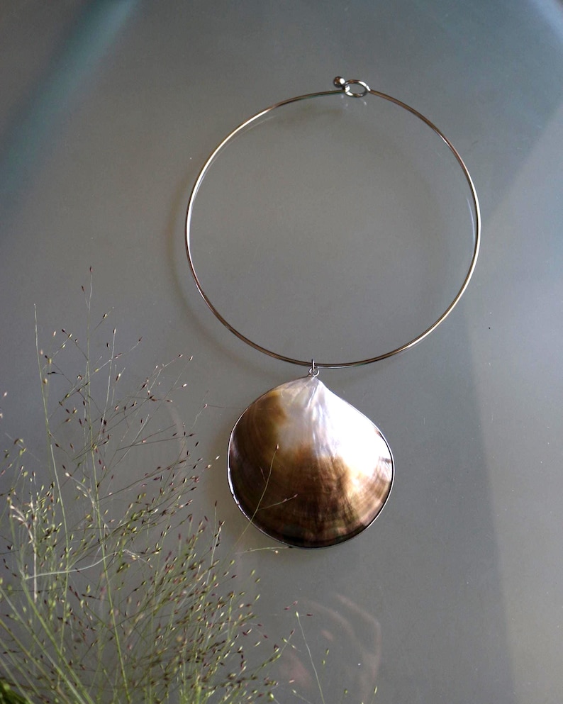 May include: A silver-toned metal choker necklace featuring a single, large, rounded seashell pendant. The shell displays a gradient of colors, transitioning from a deep, dark brown at the base to a bright, iridescent white at the top edge.