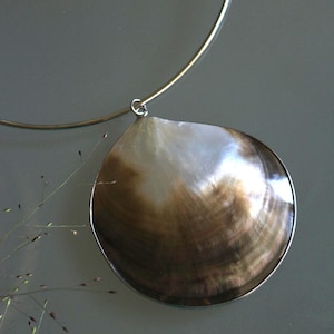 May include: A necklace featuring a large, circular, iridescent seashell pendant with a gradient of white to deep brown tones. The shell is encased in a thin silver-toned metal rim and hangs from a simple, rigid, silver-toned metal collar necklace.