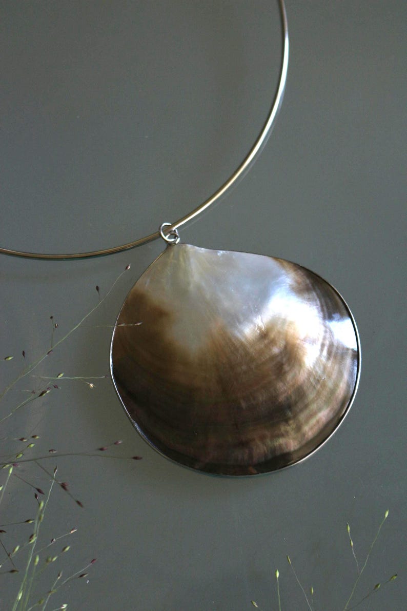 May include: A necklace featuring a large, circular, iridescent seashell pendant with a gradient of white to deep brown tones. The shell is encased in a thin silver-toned metal rim and hangs from a simple, rigid, silver-toned metal collar necklace.