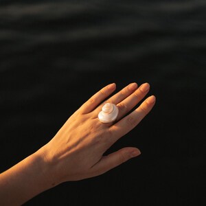 May include: A ring featuring a polished, spiraled white seashell as the central focal point. The shell has a smooth, pearlescent surface with a natural coiled structure, securely mounted on a simple, thin metallic band that encircles the finger.