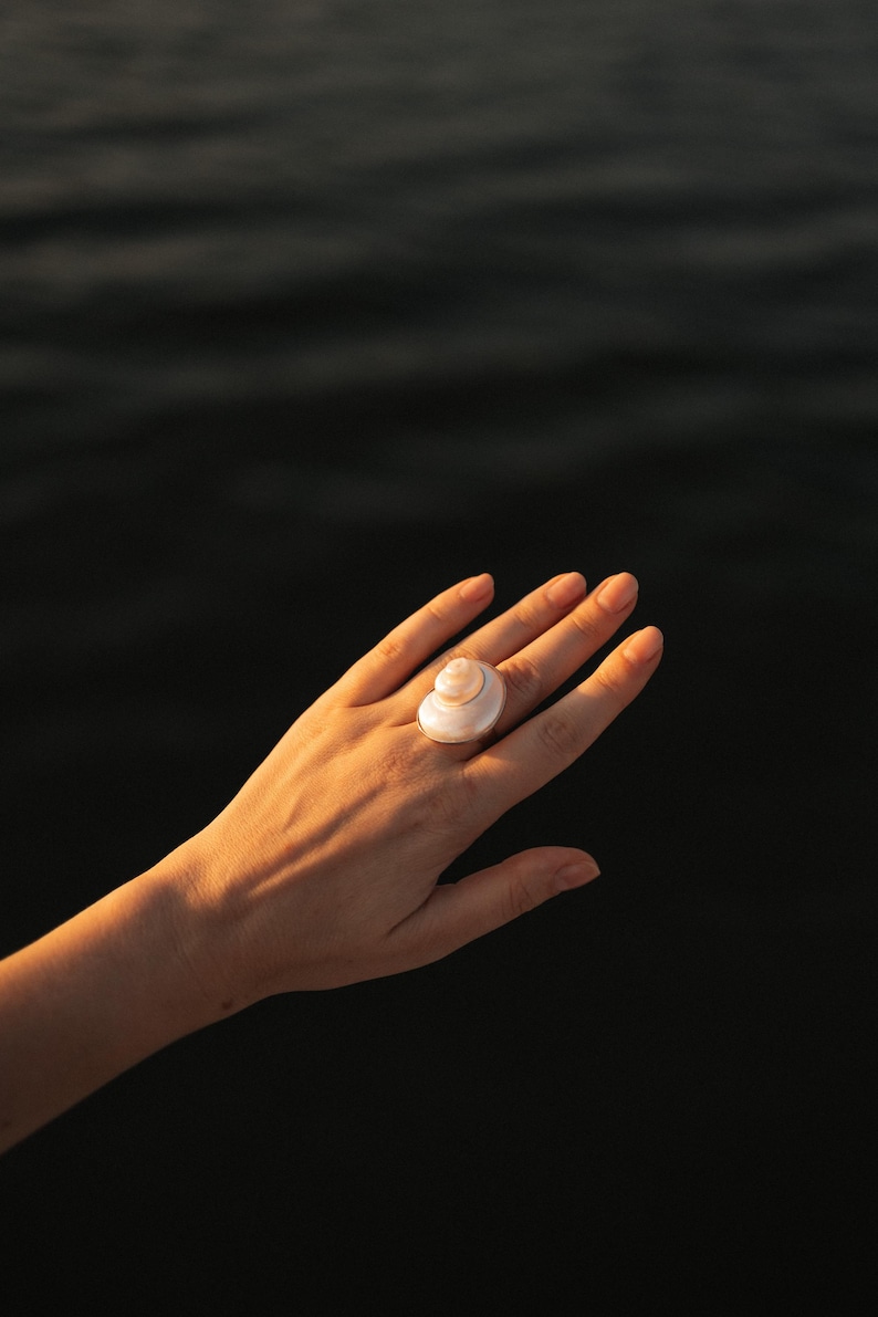 May include: A ring featuring a polished, spiraled white seashell as the central focal point. The shell has a smooth, pearlescent surface with a natural coiled structure, securely mounted on a simple, thin metallic band that encircles the finger.