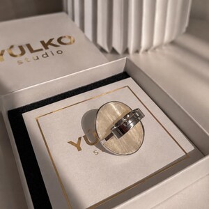 May include: A silver-toned ring with a large, oval-shaped, cream-colored inlay featuring a subtle, fibrous texture. The ring is displayed inside a white box with the text YULKO studio printed in gold on the lid and on the inner card insert.