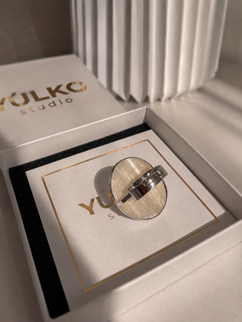 May include: A silver-toned ring with a large, oval-shaped, cream-colored inlay featuring a subtle, fibrous texture. The ring is displayed inside a white box with the text YULKO studio printed in gold on the lid and on the inner card insert.