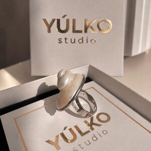 May include: A silver-toned ring featuring a polished, spiral-shaped white seashell centerpiece. The ring is displayed inside a square white box with the gold-colored text Y&Uacute;LKO studio printed on the lid and a matching vertical card behind it.