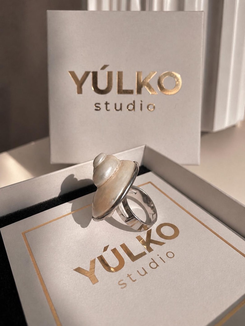 May include: A silver-toned ring featuring a polished, spiral-shaped white seashell centerpiece. The ring is displayed inside a square white box with the gold-colored text Y&Uacute;LKO studio printed on the lid and a matching vertical card behind it.