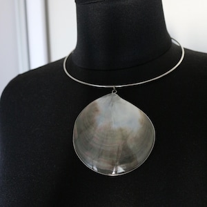 May include: A silver-toned metal choker necklace featuring a large, natural, iridescent seashell pendant. The shell displays a smooth, polished surface with swirling shades of grey, brown, and white, suspended from a thin, circular metal band.