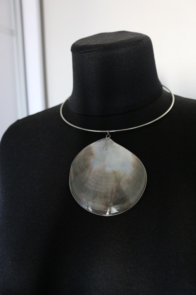May include: A silver-toned metal choker necklace featuring a large, natural, iridescent seashell pendant. The shell displays a smooth, polished surface with swirling shades of grey, brown, and white, suspended from a thin, circular metal band.