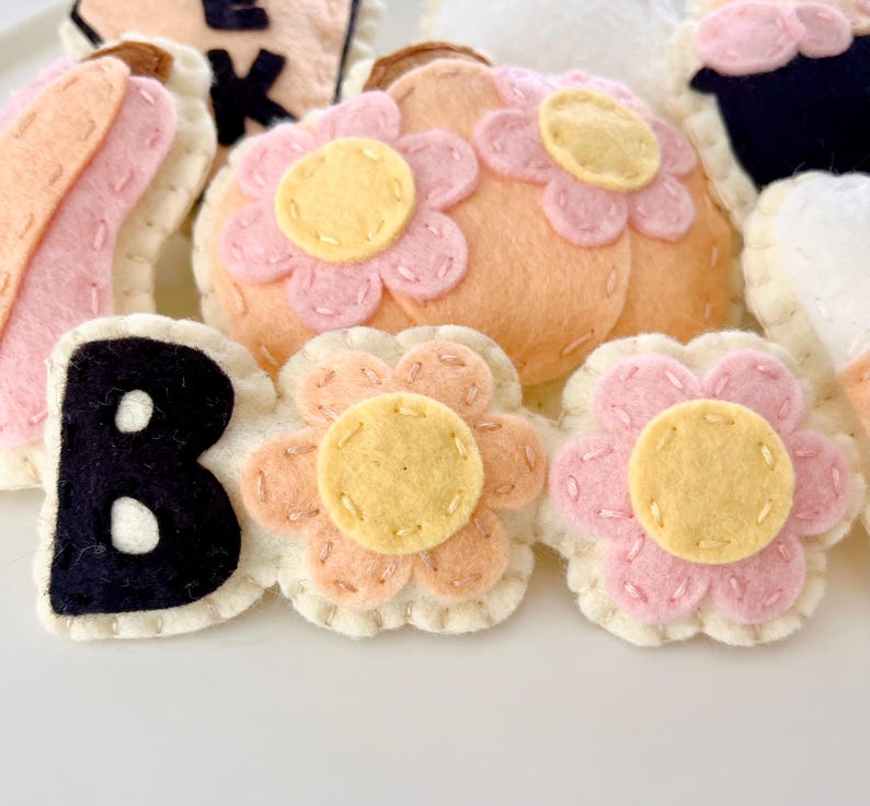 K&ouml;nnte beinhalten: Filz-Halloween-Dekorationen mit den Buchstaben "BOO" und einem K&uuml;rbis-, Bananen- und Blumendesign in Rosa, Orange und Gelb.
