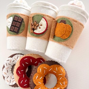 Op de afbeelding: Drie koffiebekers van vilt met verschillende herfstthema's: chocolade, appel en pompoen. Drie donuts van vilt met verschillende toppings: witte sprinkles, rode sprinkles en oranje sprinkles.