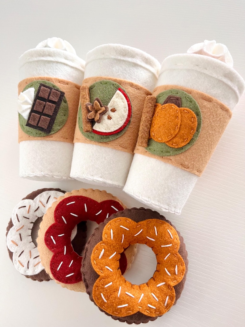 Op de afbeelding: Drie koffiebekers van vilt met verschillende herfstthema's: chocolade, appel en pompoen. Drie donuts van vilt met verschillende toppings: witte sprinkles, rode sprinkles en oranje sprinkles.