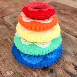 May include: A rainbow-colored felt stacking toy shaped like a donut. The toy is made of six pieces, each a different color of the rainbow, and each with a white felt circle in the center. The toy is stacked on a wooden surface.