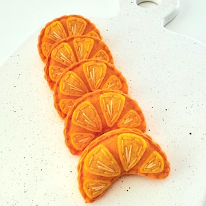 May include: Five crescent-shaped felt orange slices arranged in a vertical row. Each slice features bright orange fabric with yellow embroidered segments and light-colored stitching details, creating a textured, citrus-inspired design.